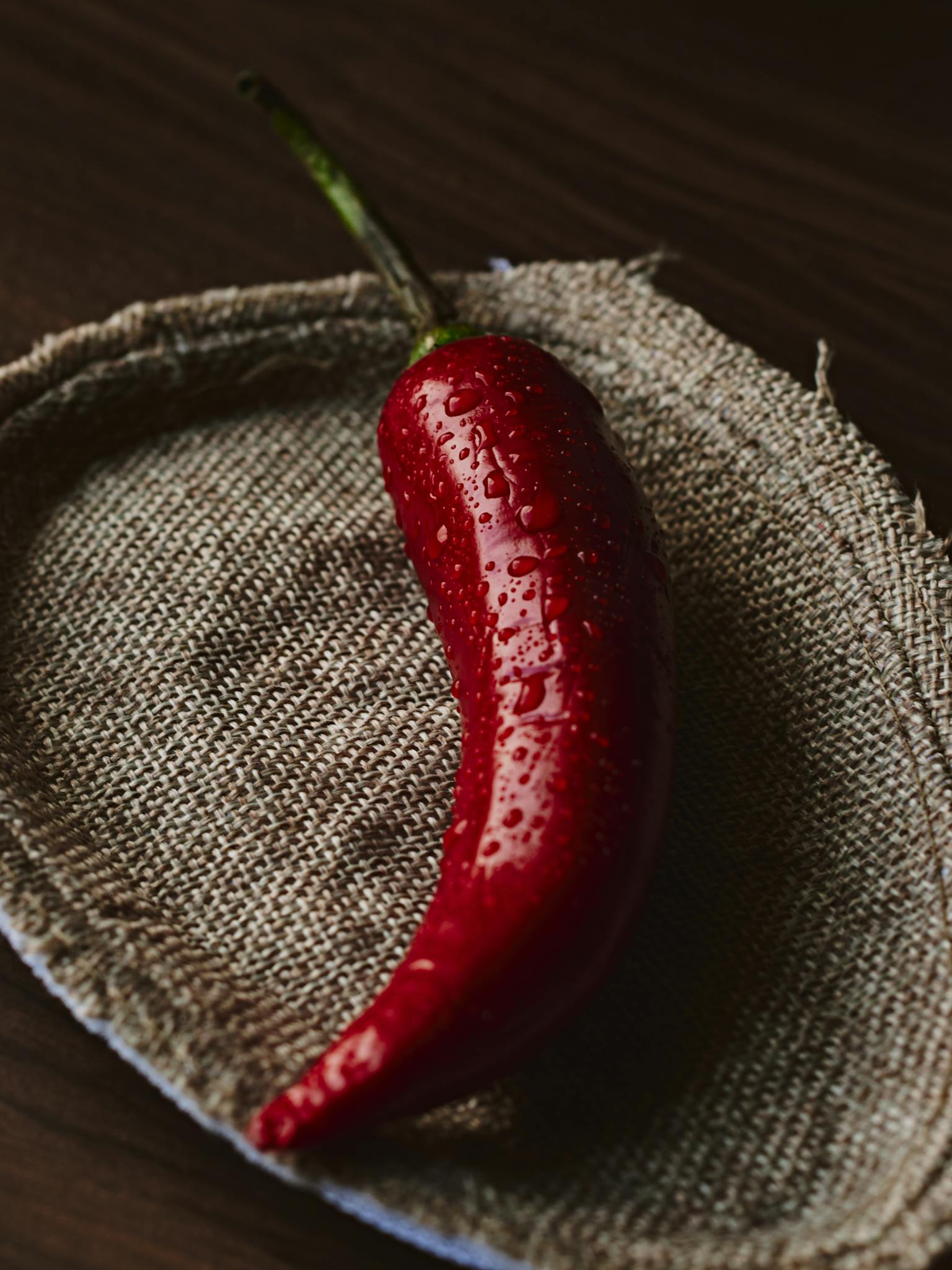 A vibrant red chili pepper with water droplets on a textured burlap surface, showcasing freshness and spice.